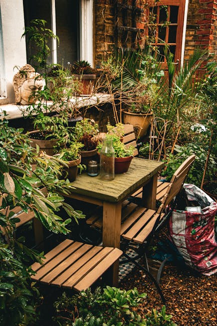 A cozy garden patio positioned against a brick house wall, featuring a weathered wooden outdoor dining table surrounded by matching wooden benches and chairs. The table is decorated with various potte